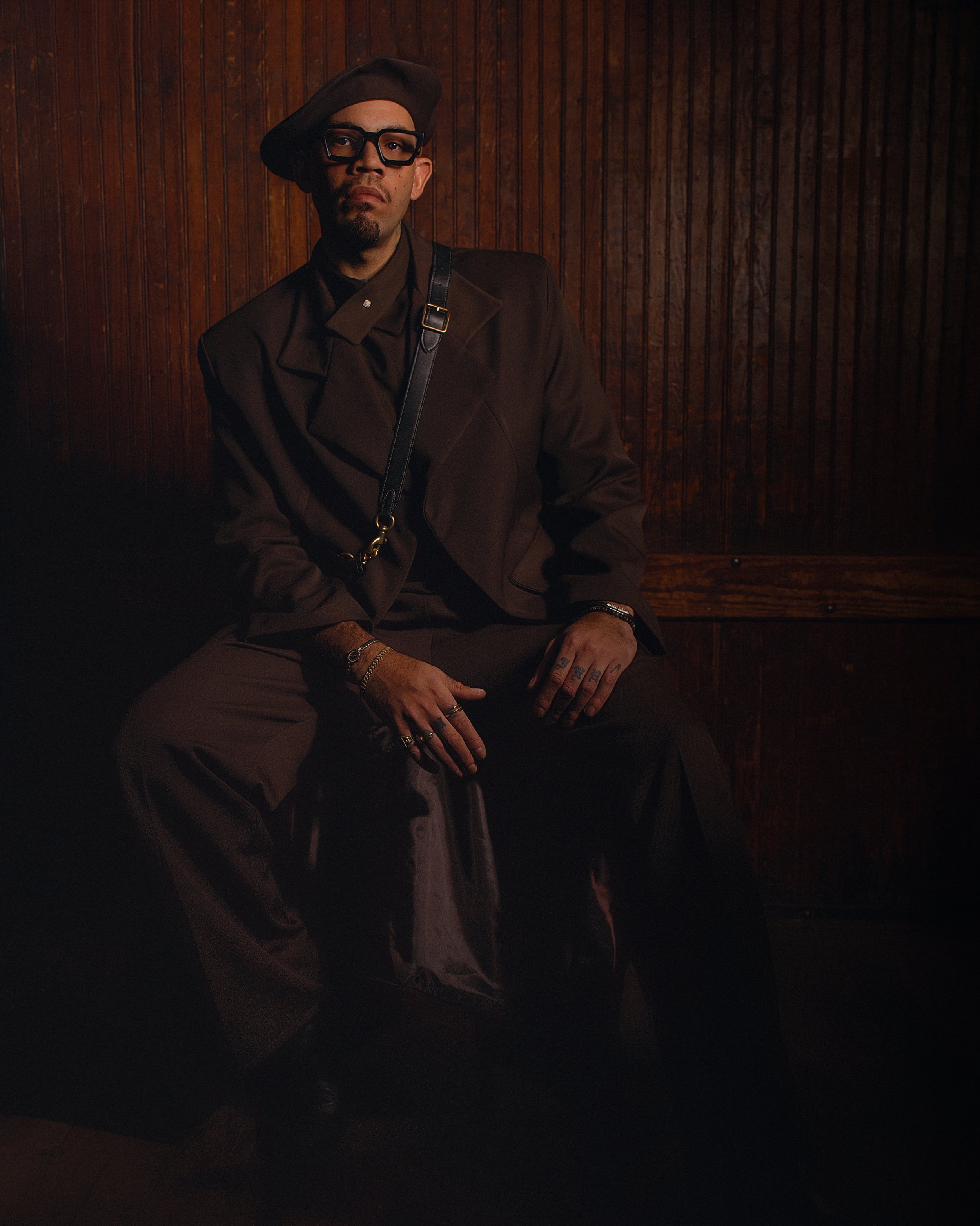 A man wearing black glasses, a black beret, a dark brown blazer, and multiple rings and bracelets sitting against a wooden paneled wall.