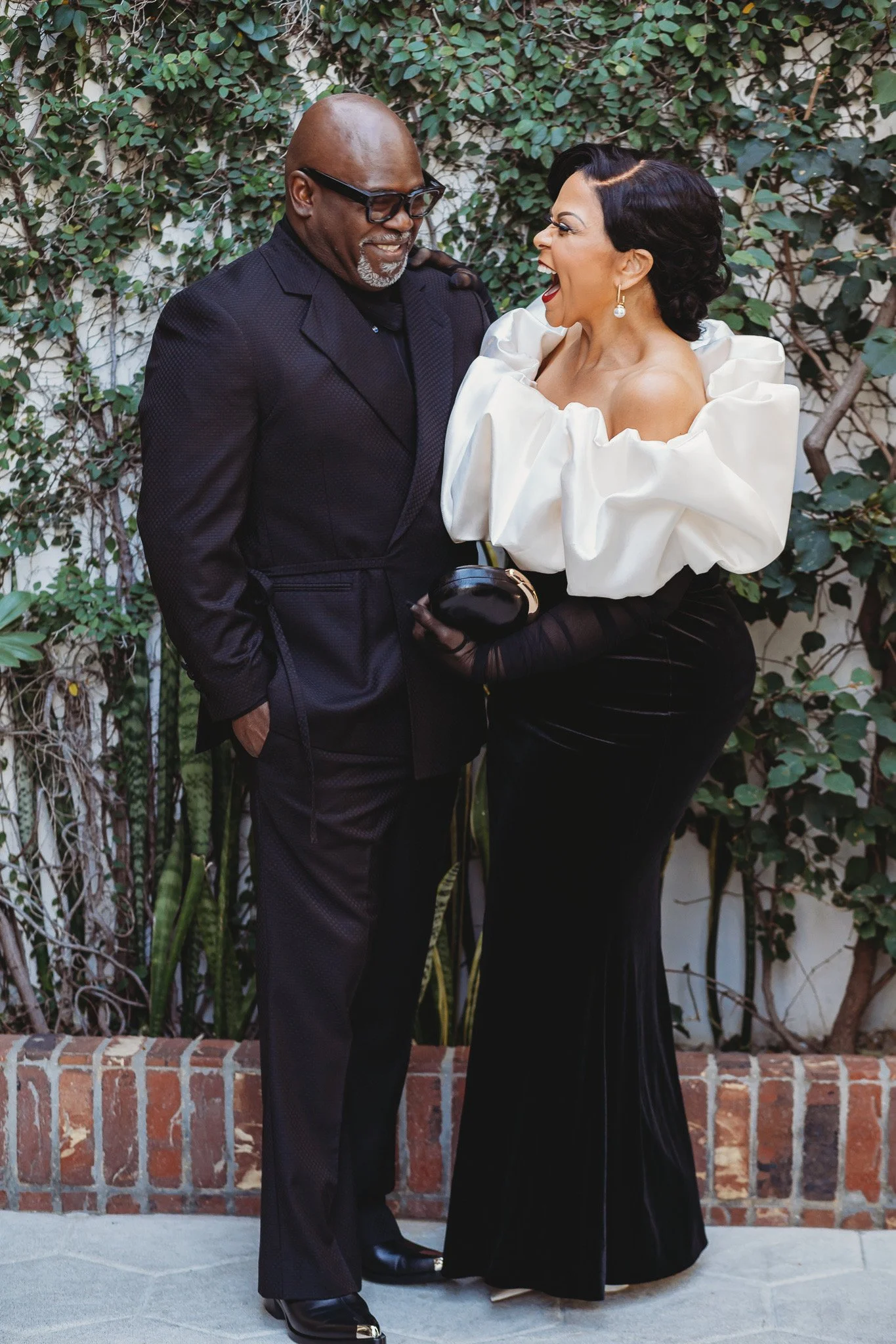 A joyful woman and man are laughing and smiling together outdoors, standing close in front of a green vine-covered wall.