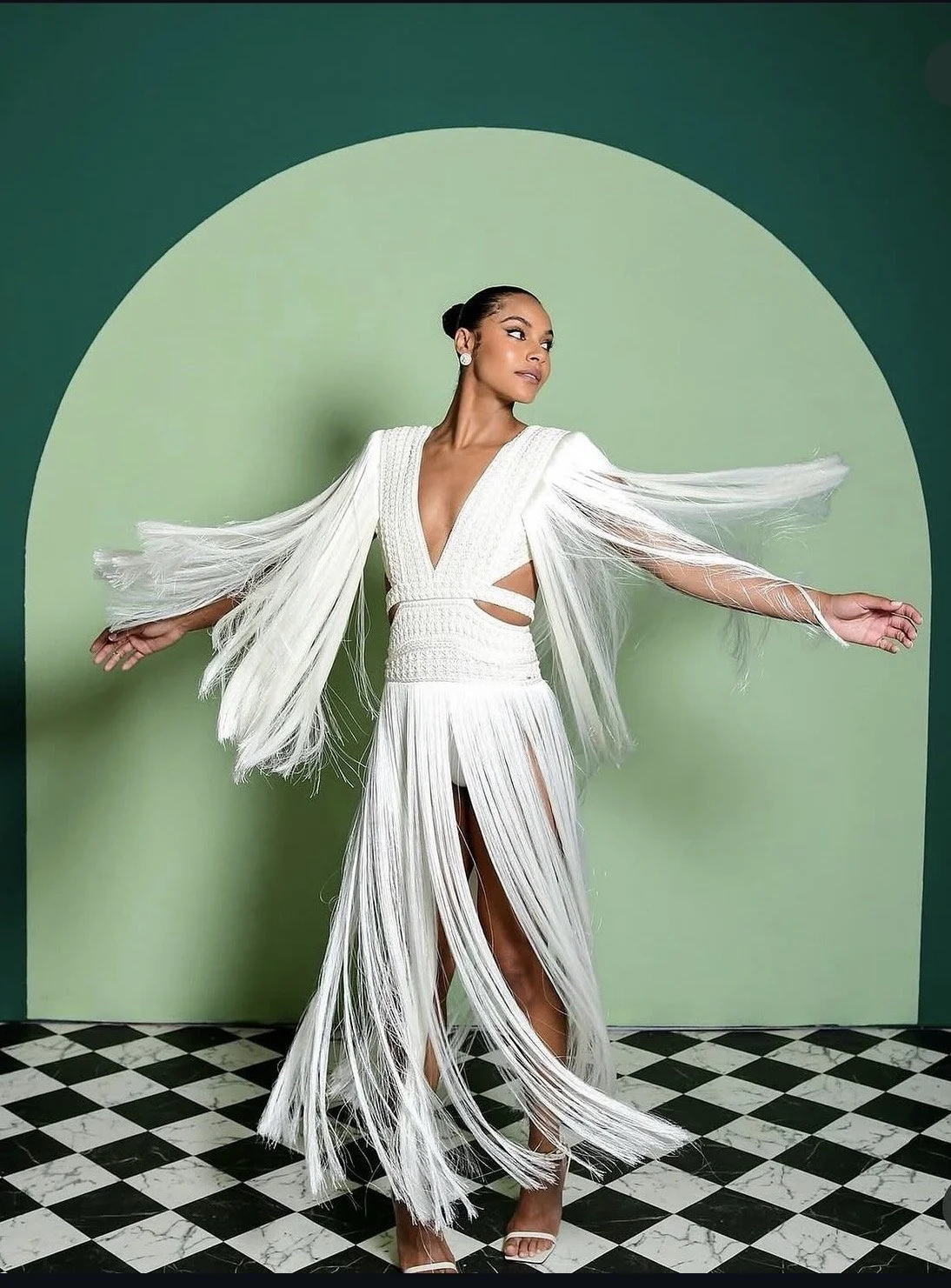 A woman in a white, fringed dress with a deep V-neckline and cutouts, standing against a green background with an arch, on a black and white checkered floor.