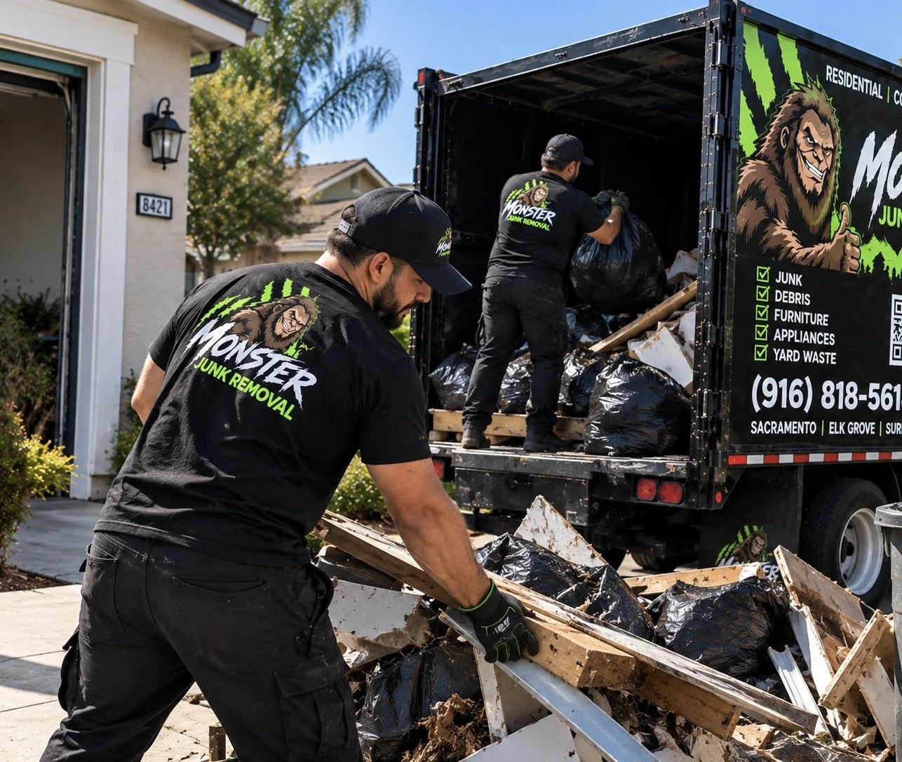 Men wearing black shirts and hats with 'Monster Junk Removal' logo load debris and trash into a large black truck with similar branding on the side, parked outside a house.
