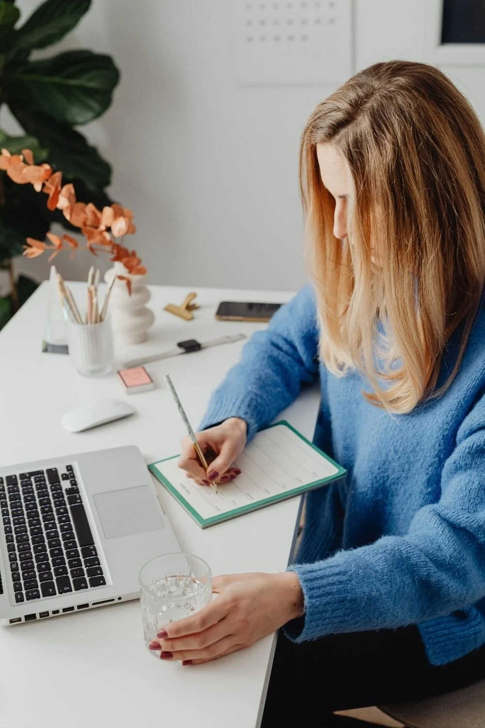 Woman in a blue sweater writing in a planner at a desk with a laptop and glass of water