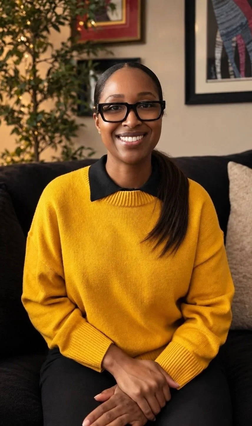 Smiling woman wearing black glasses and a mustard yellow sweater seated on a sofa