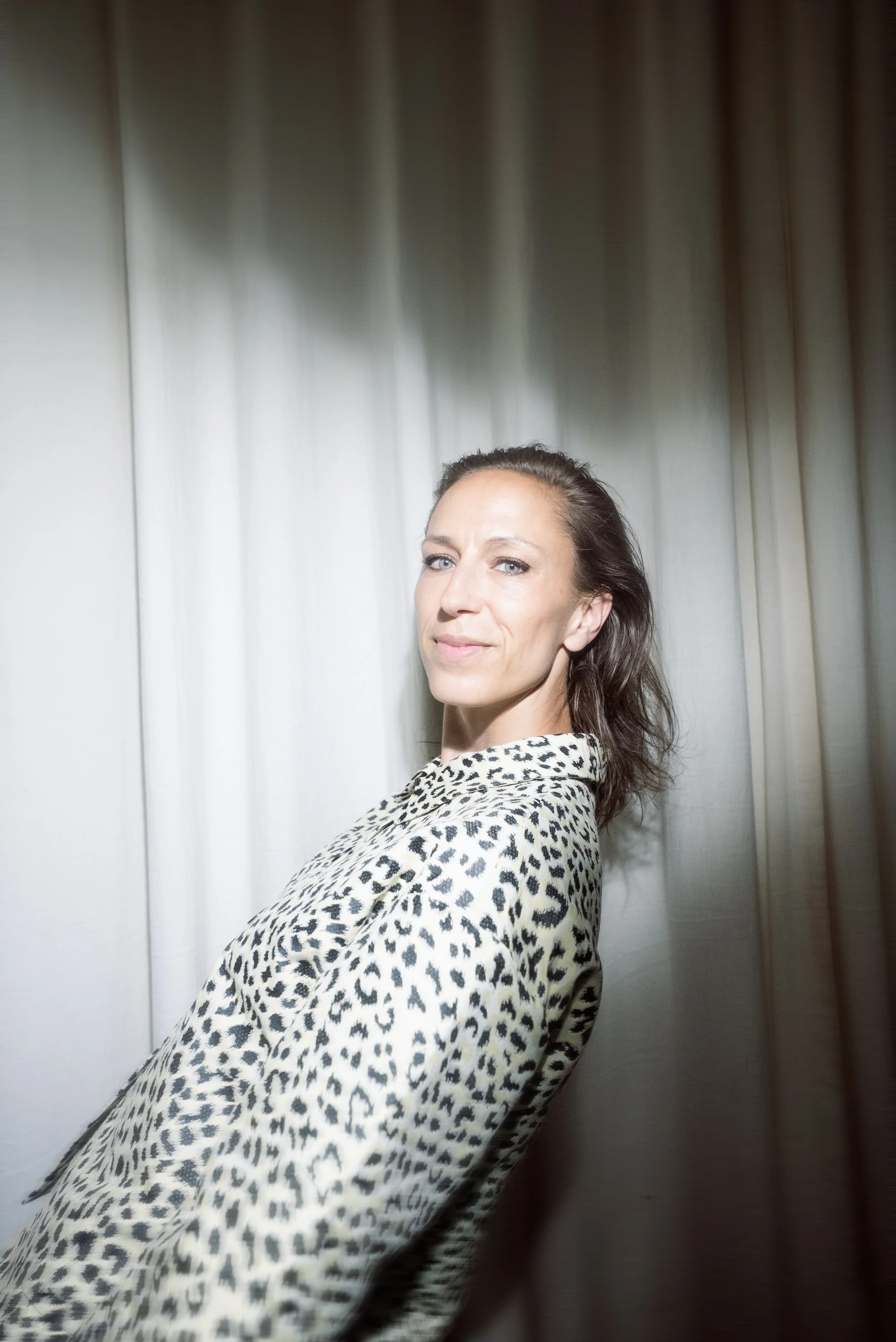 Portræt af en kvinde med langt mørkt hår, iført leopardmønstret bluse, stående foran en lys væg med gardiner.