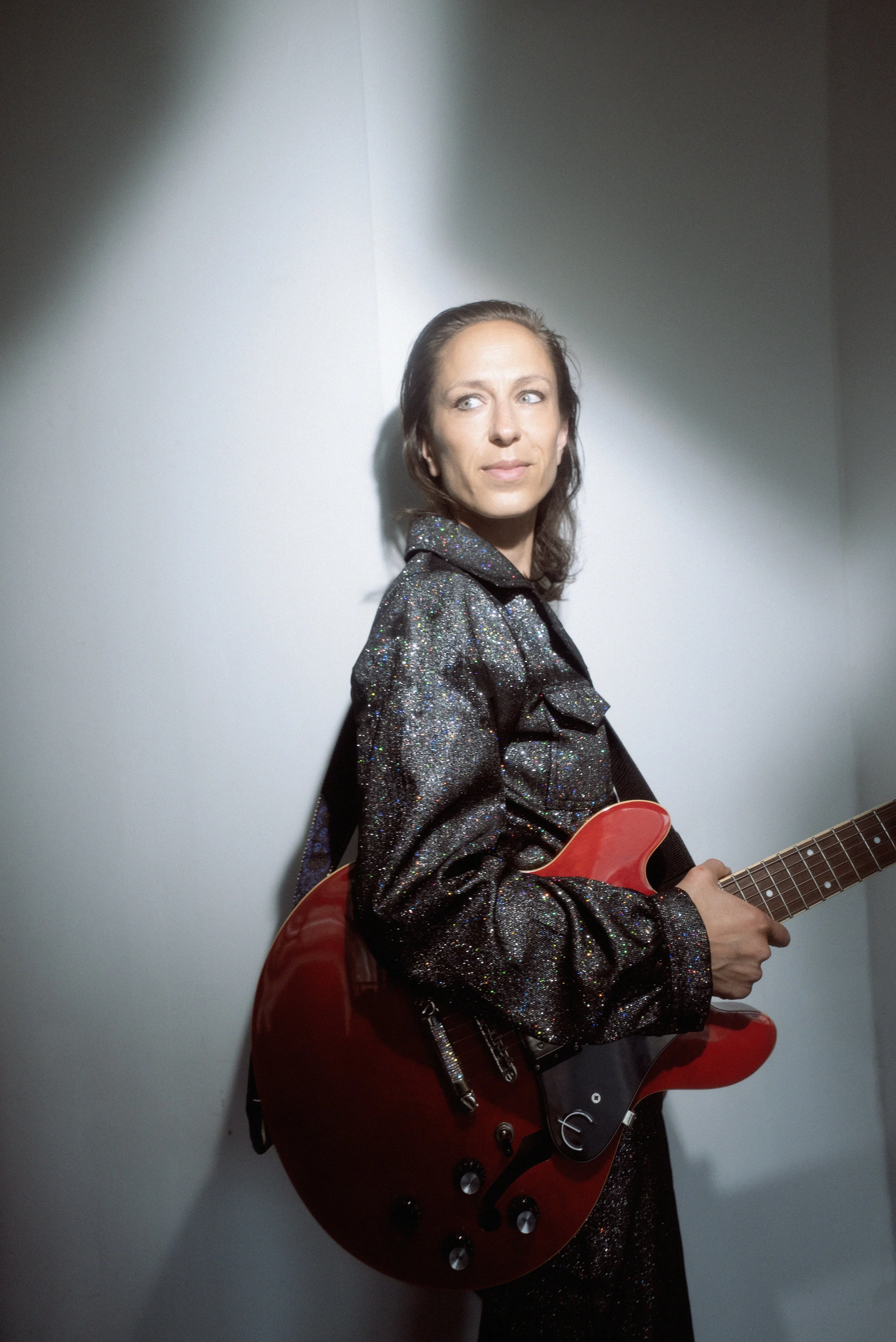 Kvindelig musiker med langt mørkt hår, iført en glitrende sort jakke, spiller på en rød elektrisk guitar, står mod en hvid væg under belysning.
