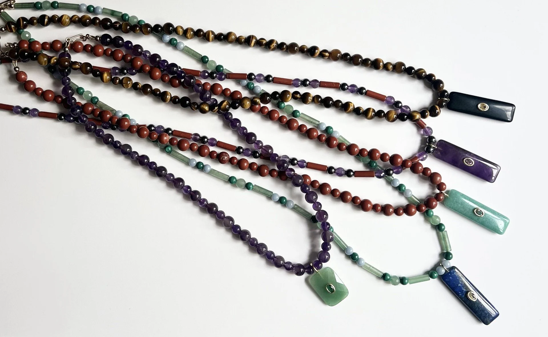 Multiple beaded necklaces with colourful semi-precious beads and rectangular semi precious pendants on a white background.