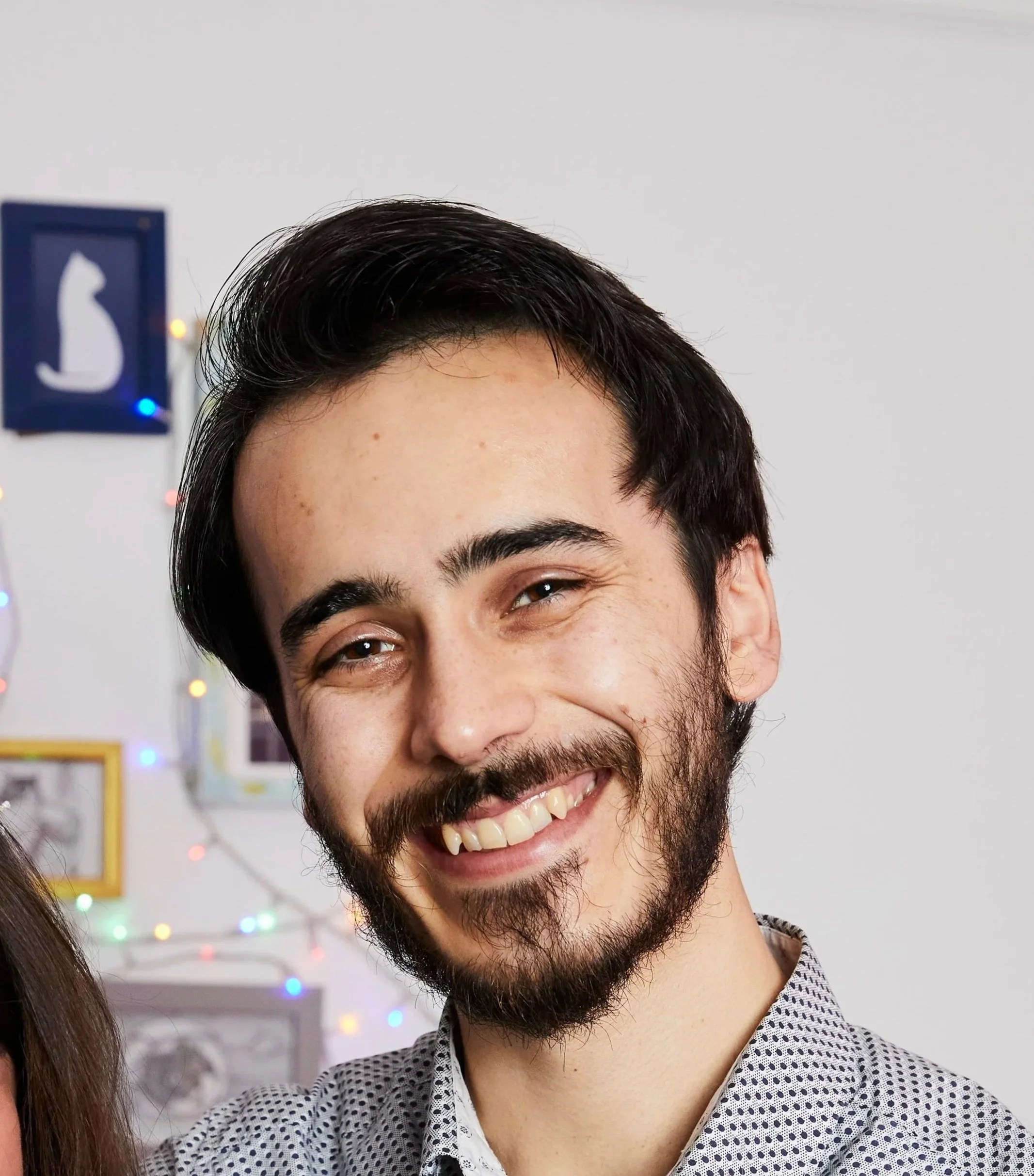 Close-up of a smiling young man with dark hair and a beard, wearing a polka dot shirt, standing in front of a white wall decorated with colorful string lights.
