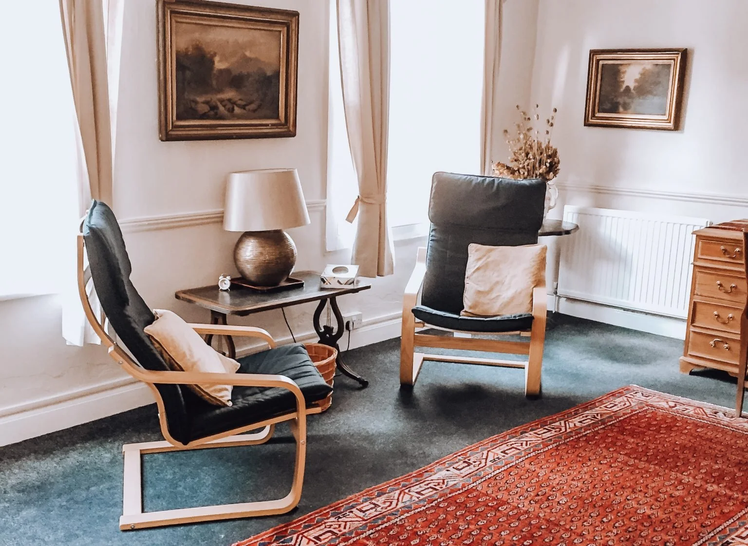 A cozy corner of a counselling room with two black armchairs with wooden frames, a side table with a lamp, artworks on the wall, a window with curtains, a patterned rug, and a dresser with a vase of dried flowers.