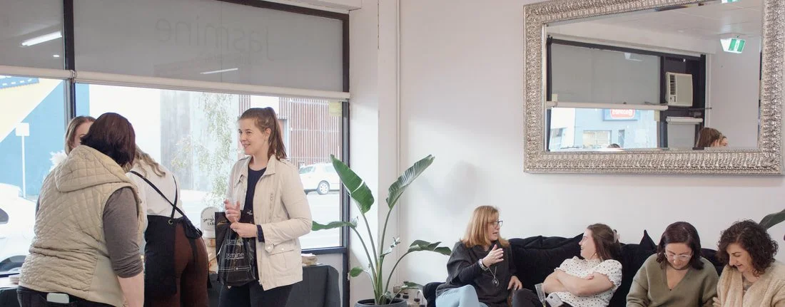 Group of women socializing indoors, some seated on black sofa, some standing near window, in a room with large mirror and indoor plants.