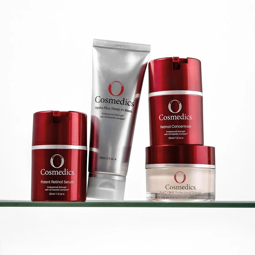Collection of OCosmedics skincare products including retinol serum, sleep-in mask, retinol concentrate, and a cream in red and white packaging on a glass shelf.