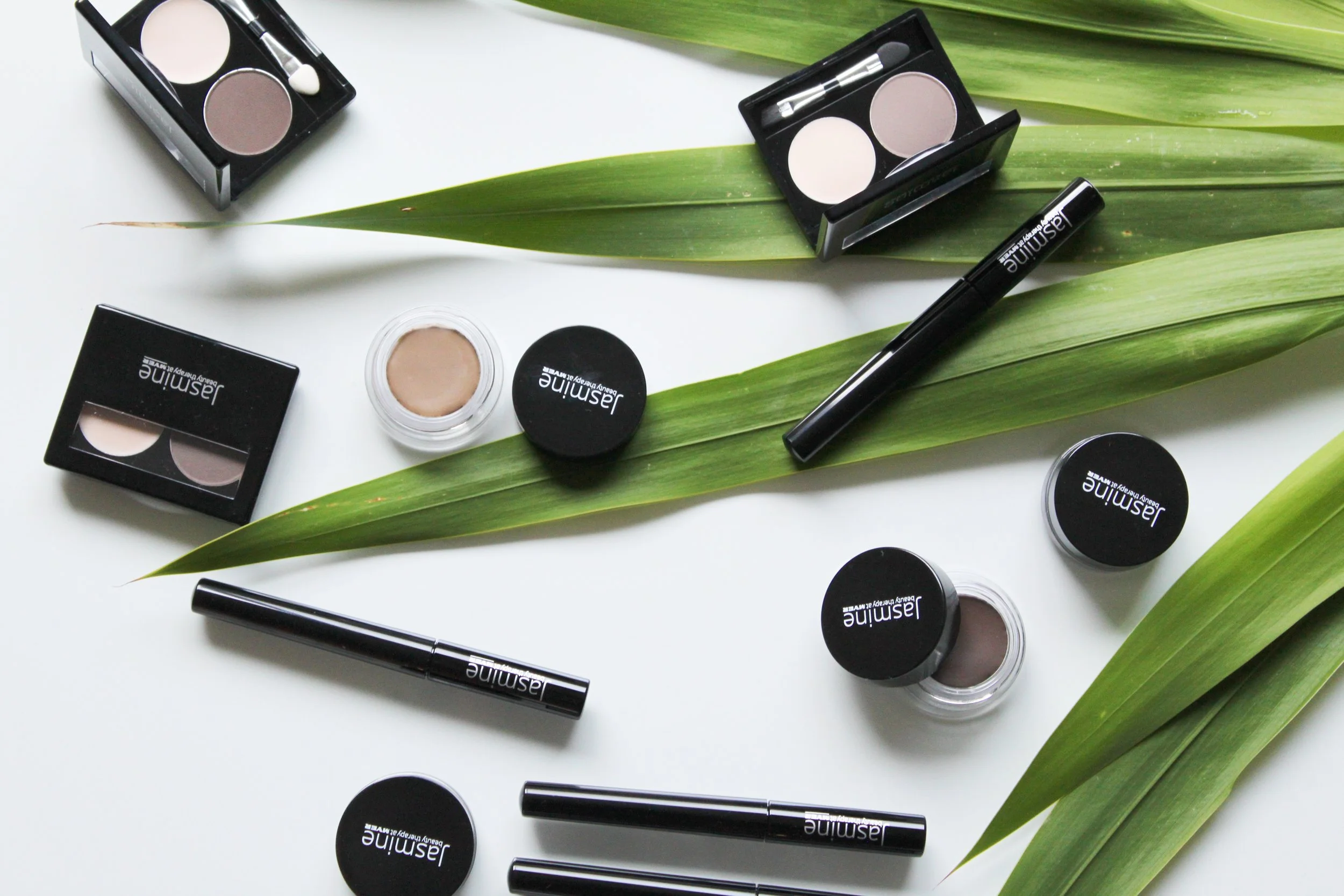 Makeup products for eyebrows, including a brow pencil and brow fixx mascara, arranged on a white surface with large green leaves.