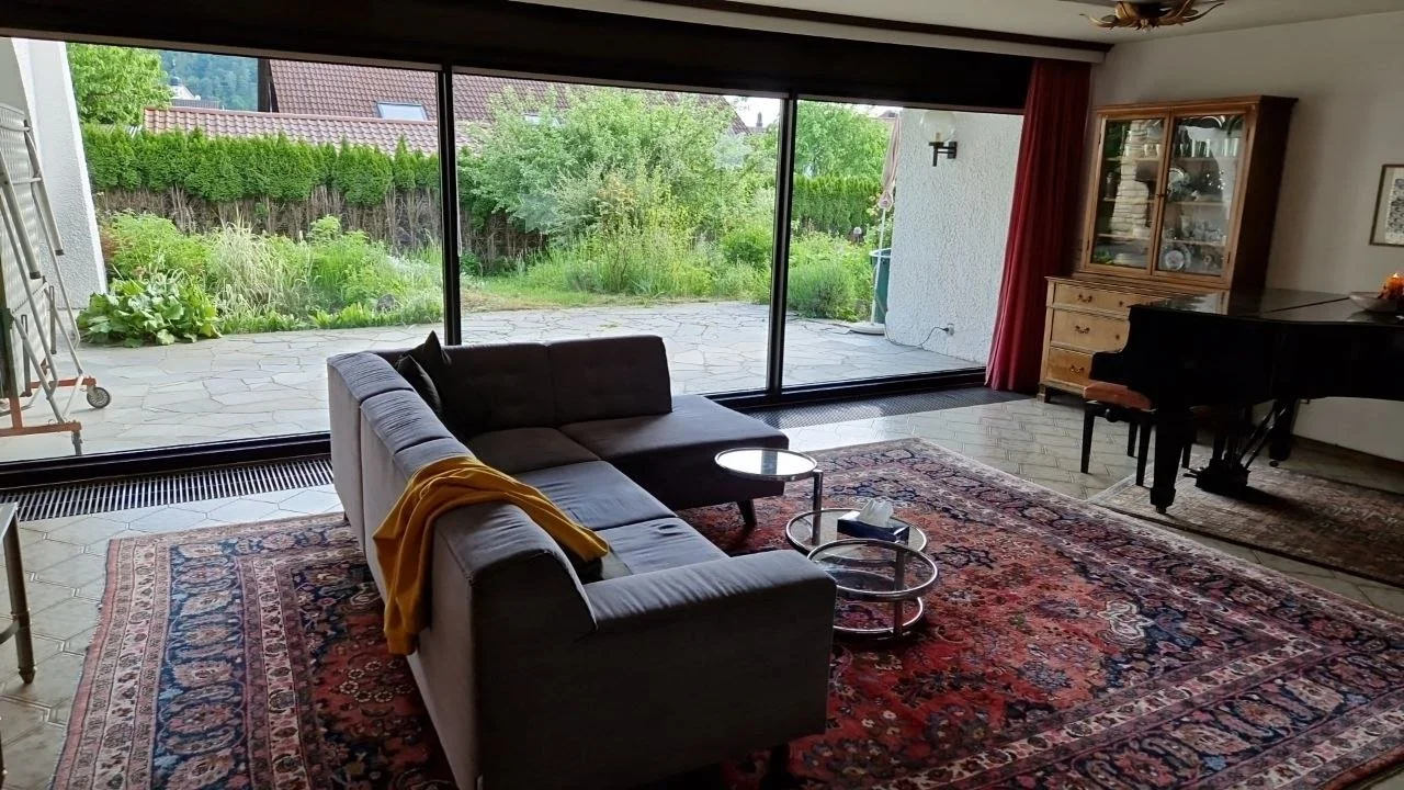 Wohnzimmer mit Blick auf den Garten, graues Sofa, Teppich, Klavier, Glastür zum Garten, Holzschrank, kleine Tische, Gardinen, Wanddekoration, Pflanzen draußen.
