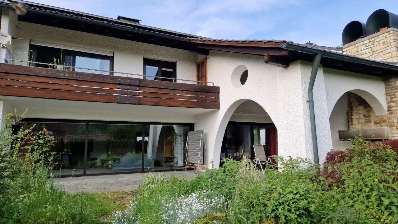 Ein modernes Haus mit weißer Fassade, großen Glasflächen und einem Balkon mit Holzgitter. Im Vordergrund befindet sich ein Garten mit verschiedenen Pflanzen. Es ist ein sonniger Tag.