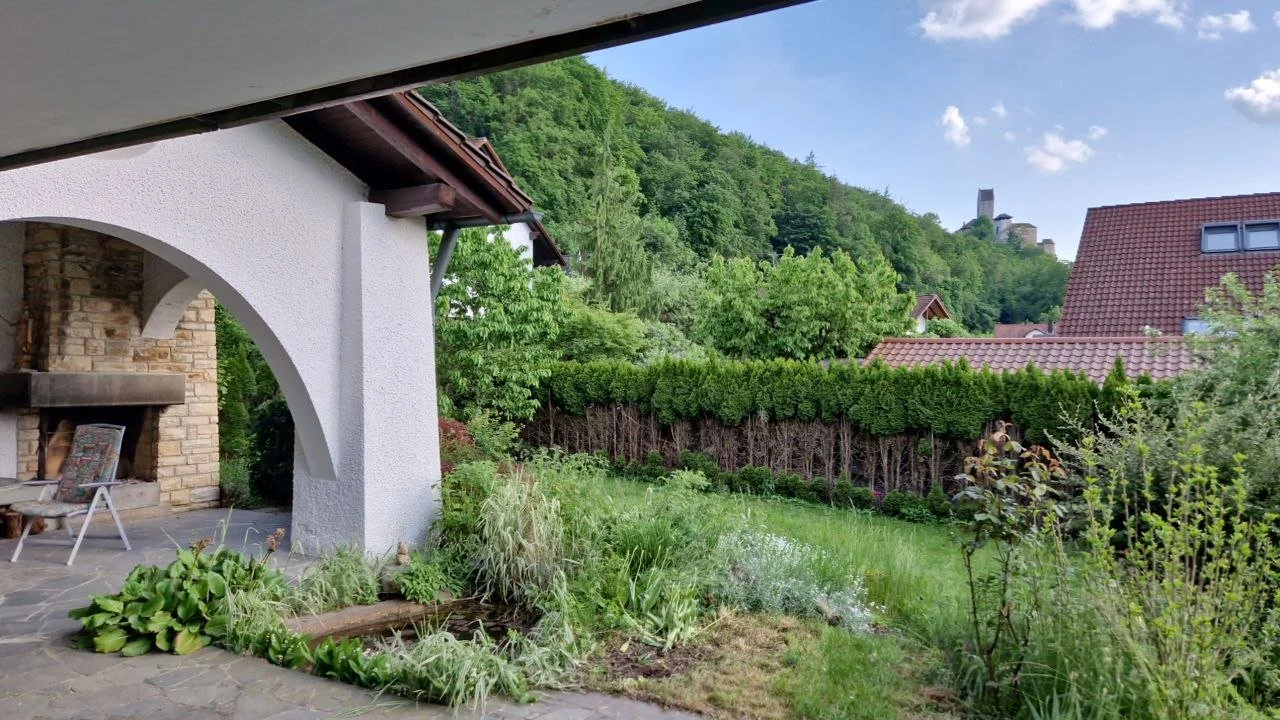 Garten mit Rasen, Sträuchern und Bäumen neben einem weißen Haus mit Terrasse, Backstein Kamin im Garten, Berge im Hintergrund, blauer Himmel mit Wolken.