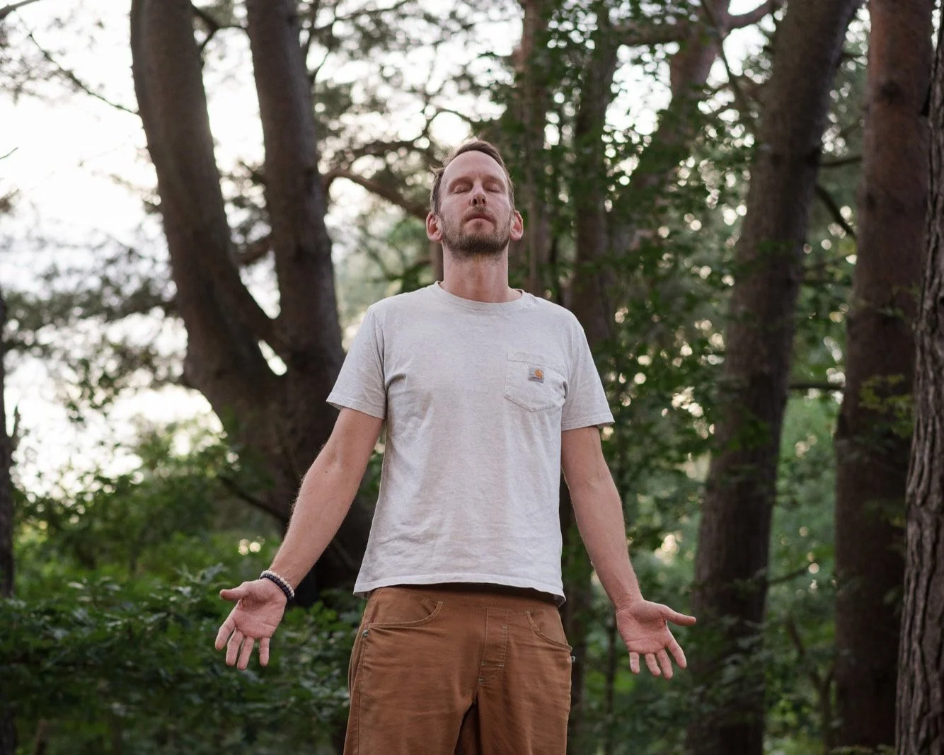 Ein Mann steht entspannt in einem Wald mit geschlossenen Augen und leicht geöffnetem Mund, die Arme seitlich ausgestreckt, in einer meditativen Haltung.