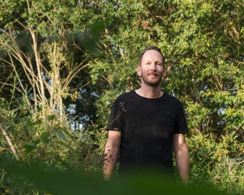 Ein Mann mit dunklem T-Shirt steht in einem grünen, bewachsenen Wald oder Park, umgeben von Bäumen und Pflanzen, bei Sonnenlicht.