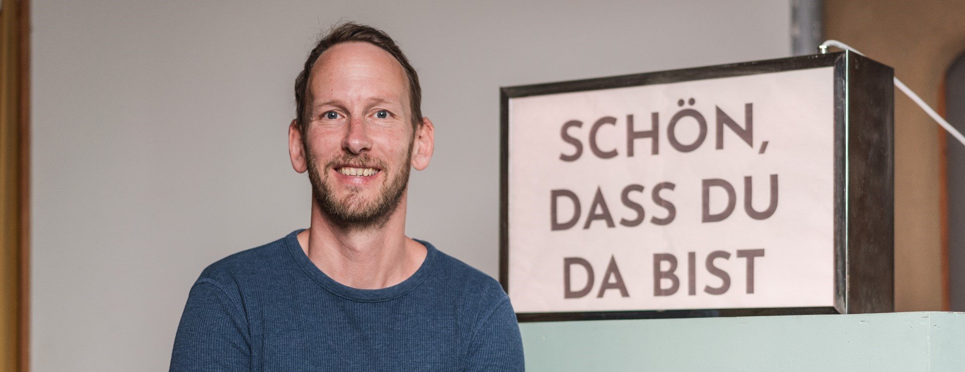 Ein lächelnder Mann mit braunem Bart und blauen Pullover steht vor einem Schild, das auf Deutsch 'Schön, dass du da bist' sagt.