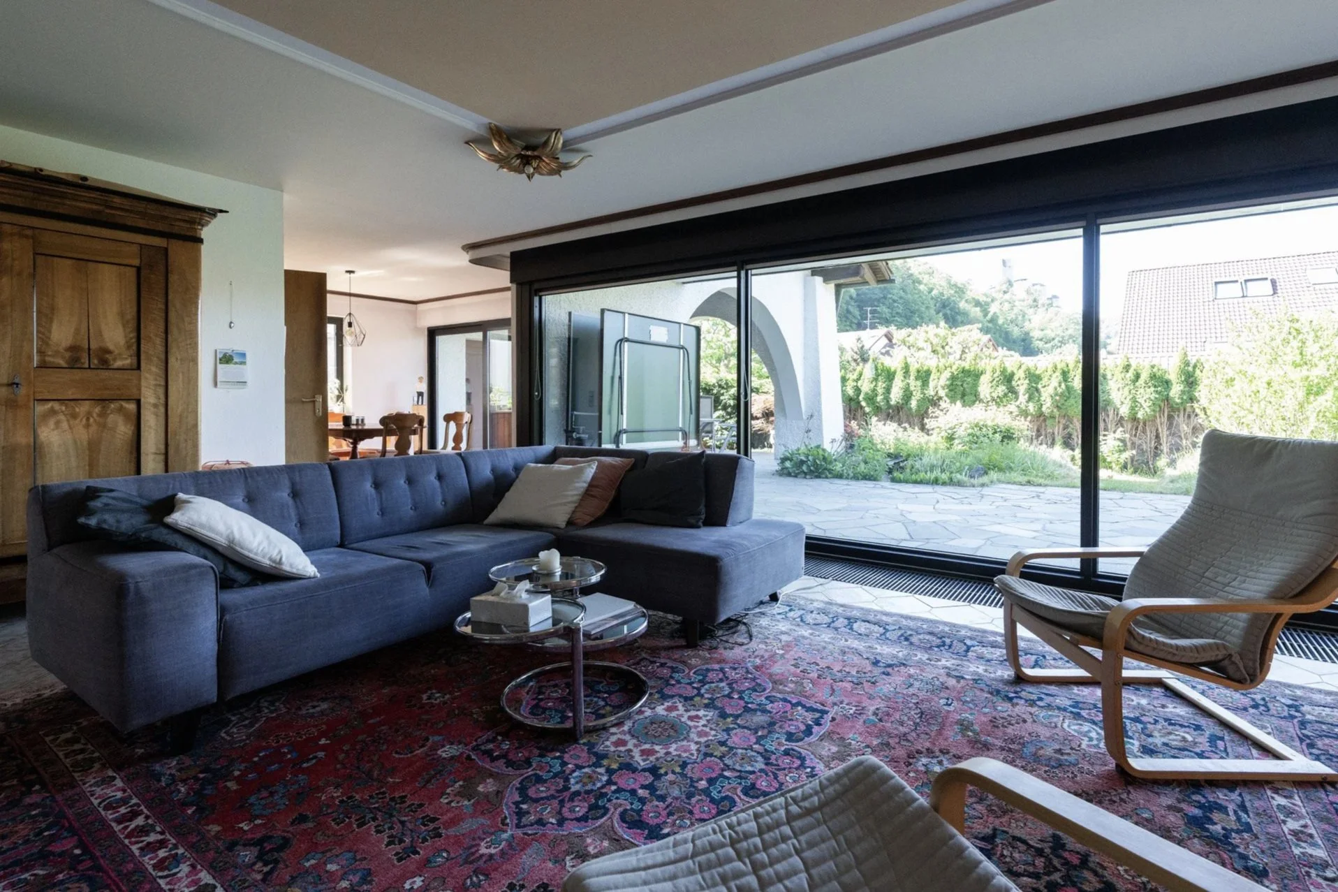 Innenansicht eines Wohnzimmers mit dunkelblauem Sofa, Kissen, einem kleinen Couchtisch, einer antiken Teppich und großen Fenstern, die einen Blick auf einen Garten bieten.