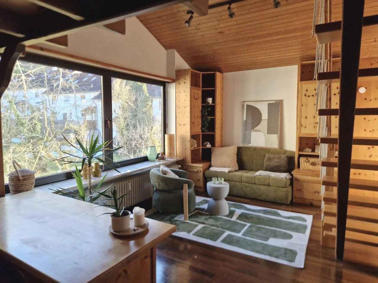 Gemütliches Wohnzimmer mit großen Fenstern, grüner Couch, Sessel, kleinen Beistelltisch, Holzregalen und moderner Wanddekoration, natürlicher Lichtquelle.
