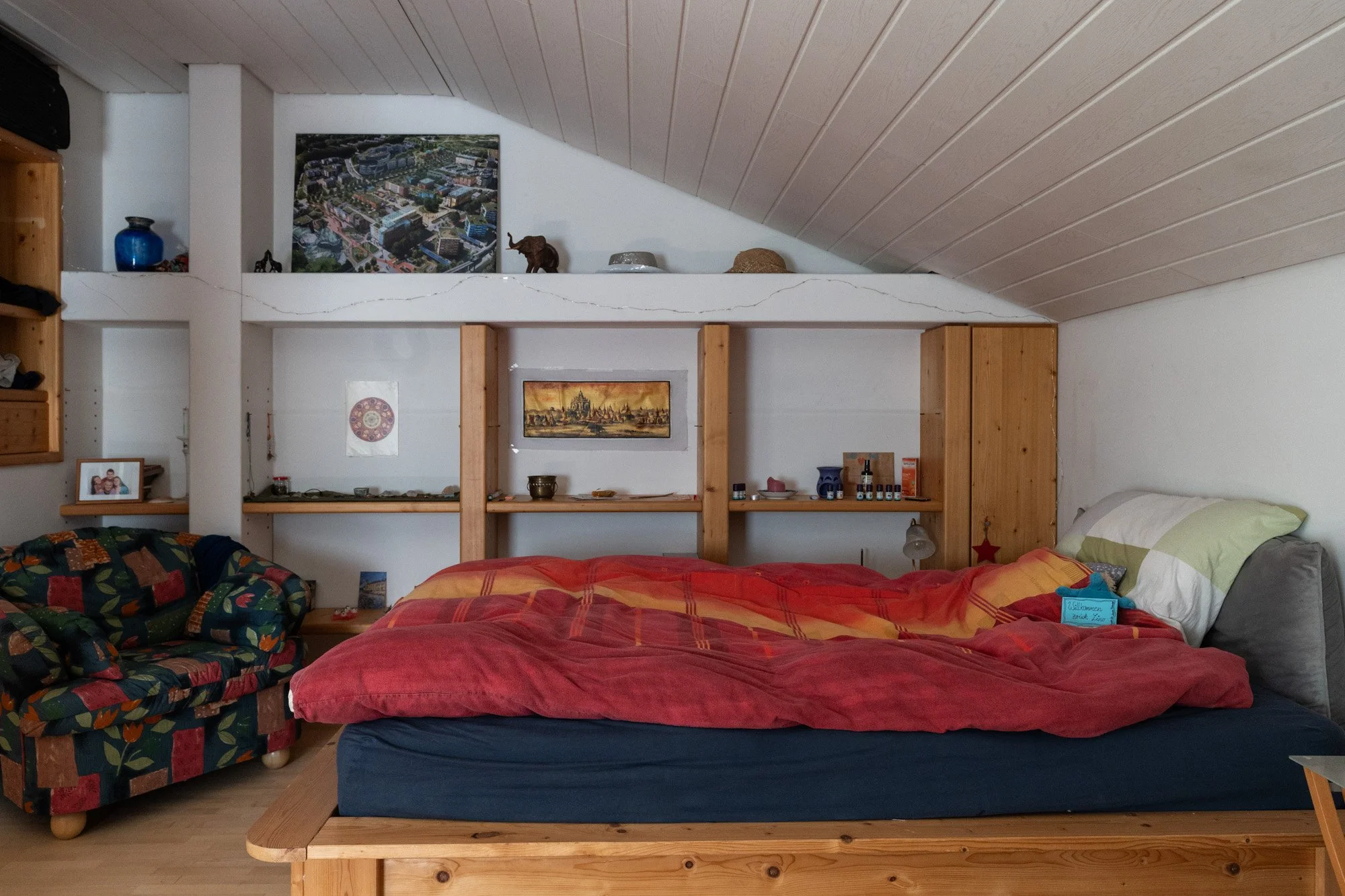 Blick in ein Schlafzimmer auf einem Bett mit roten Decken, eine bunte Couch, Wandregale mit Dekorationen und Bildern, auf dem Dachgeschoss mit Leseecke.