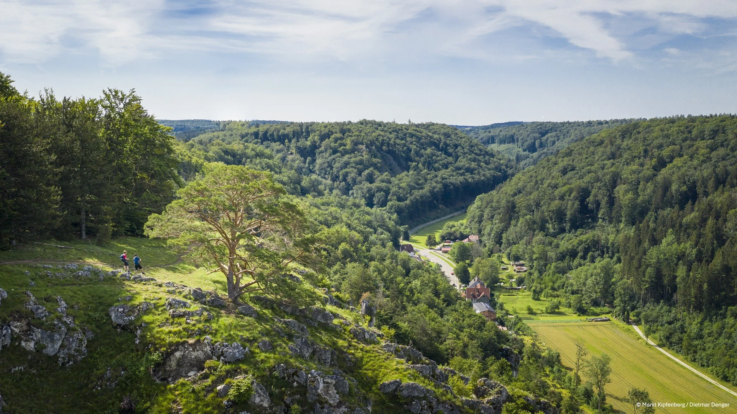 Hügelige, bewaldete Landschaft mit einem Weg, kleinen Gebäuden und zwei Personen beim Wandern.