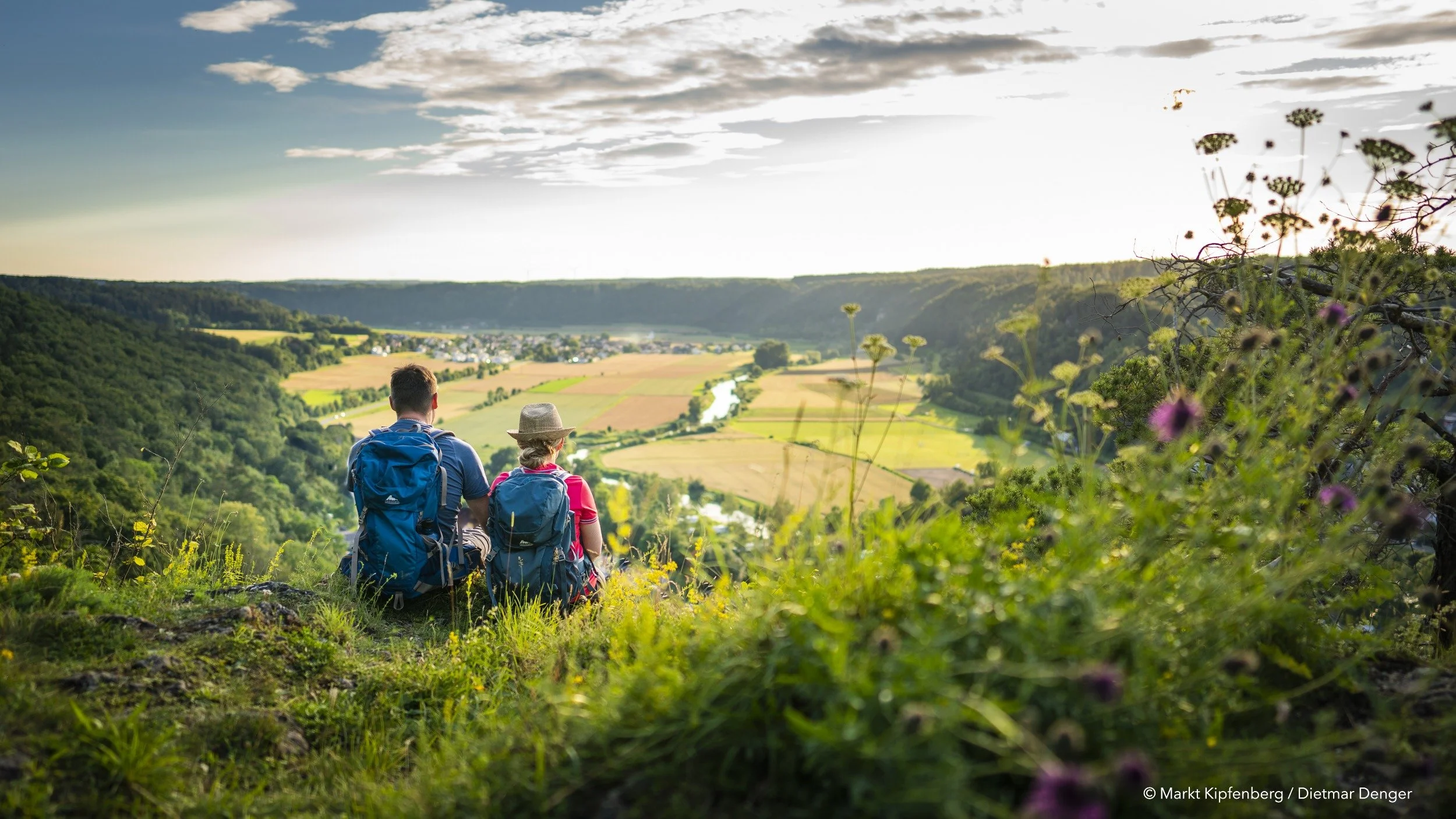 Zwei Wanderer mit Rucksäcken sitzen auf einem Hügel und schauen auf eine grüne Landschaft mit Fluss, Feldern und Dörfern bei Sonnenuntergang.