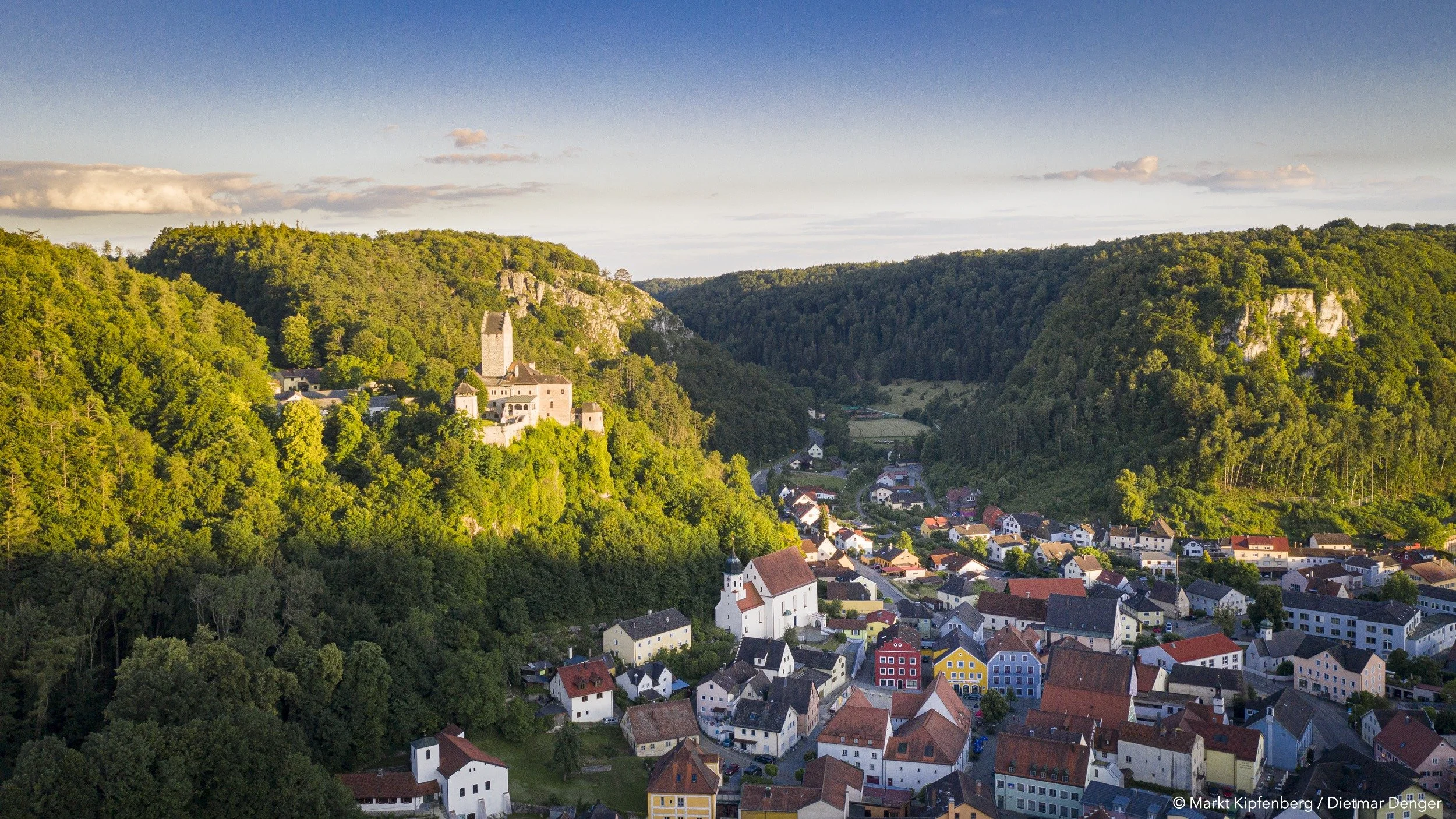 Blick auf eine kleine Stadt in einer waldreichen Hügellandschaft bei Sonnenuntergang, mit historischen Gebäuden, Kirche und bunter Wohnhäusern