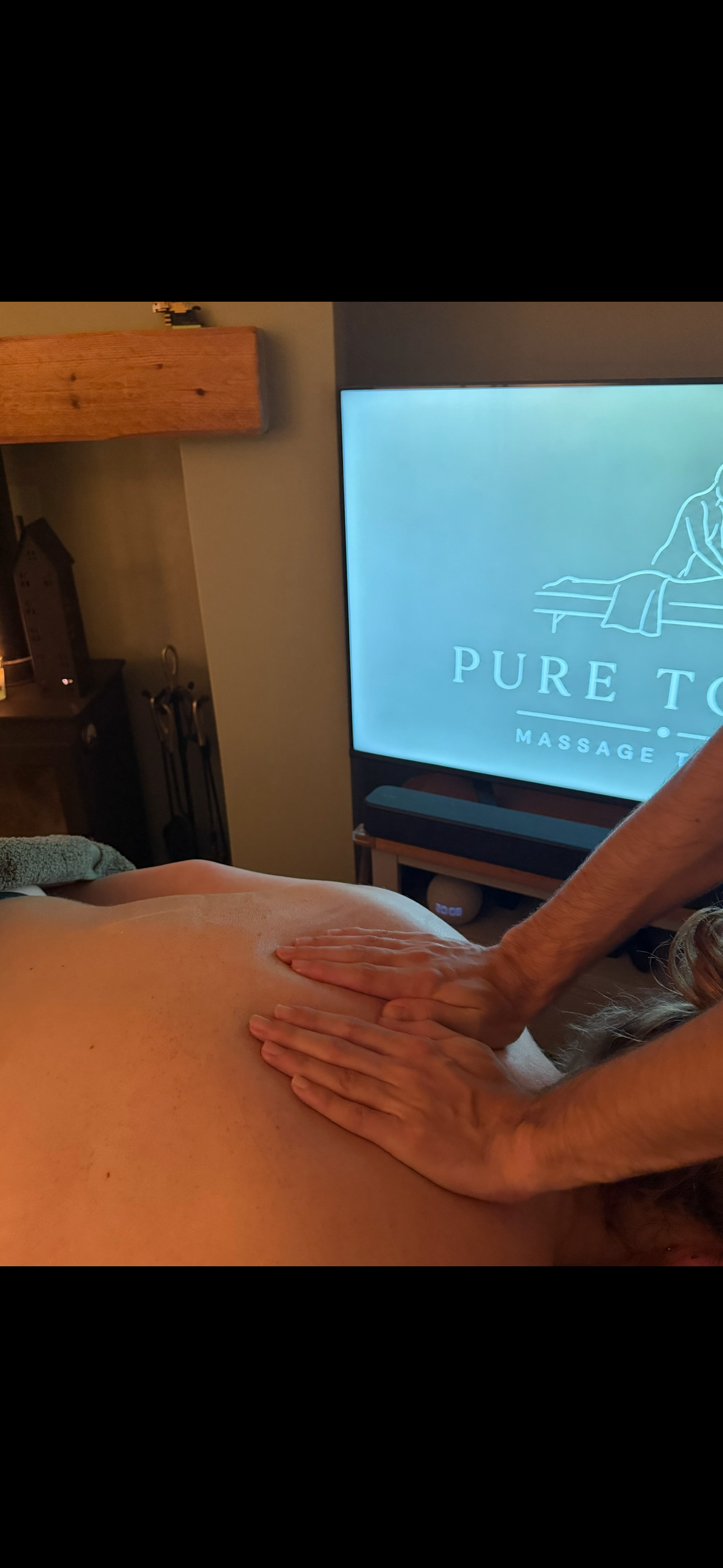 Massage therapist performing a back massage on a client in a home treatment room, with a Pure Touch Massage TV display in the background.