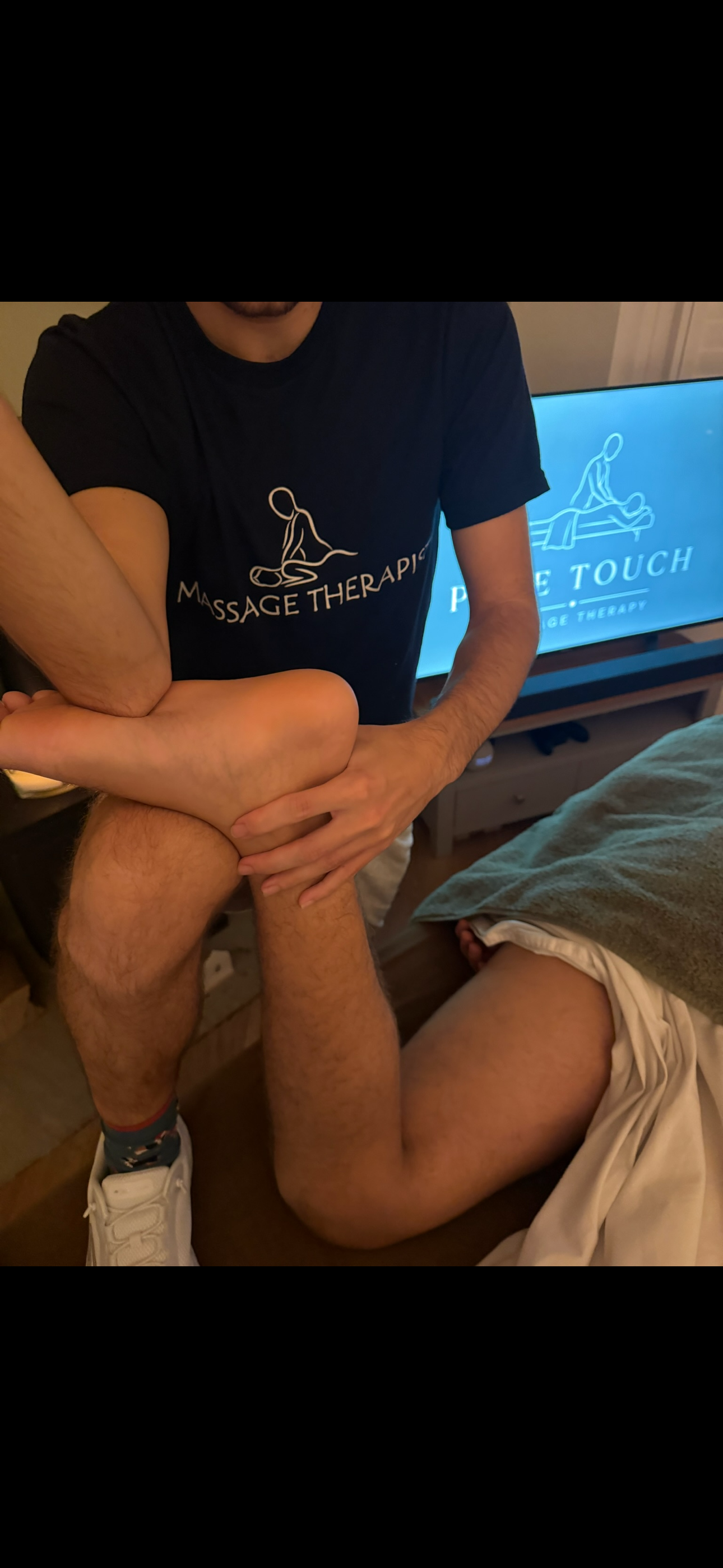 Massage therapist in a branded shirt performs a seated leg stretch on a client beside a massage therapy sign.