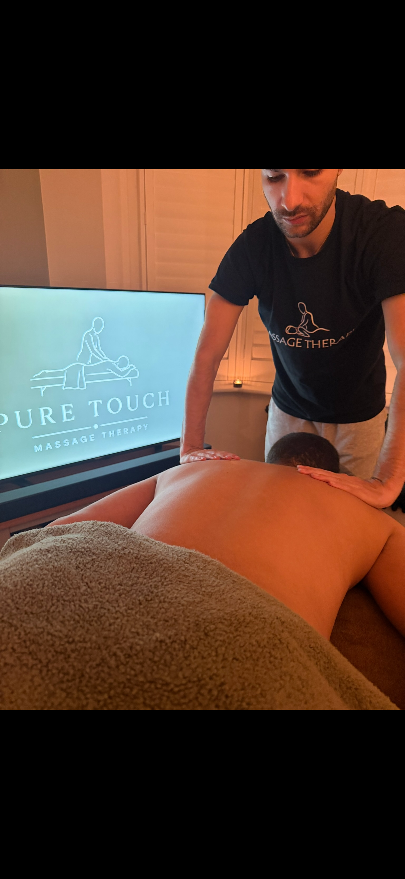 Massage therapist providing a back massage to a client in a warmly lit spa, with a Pure Touch Massage Therapy sign in the background