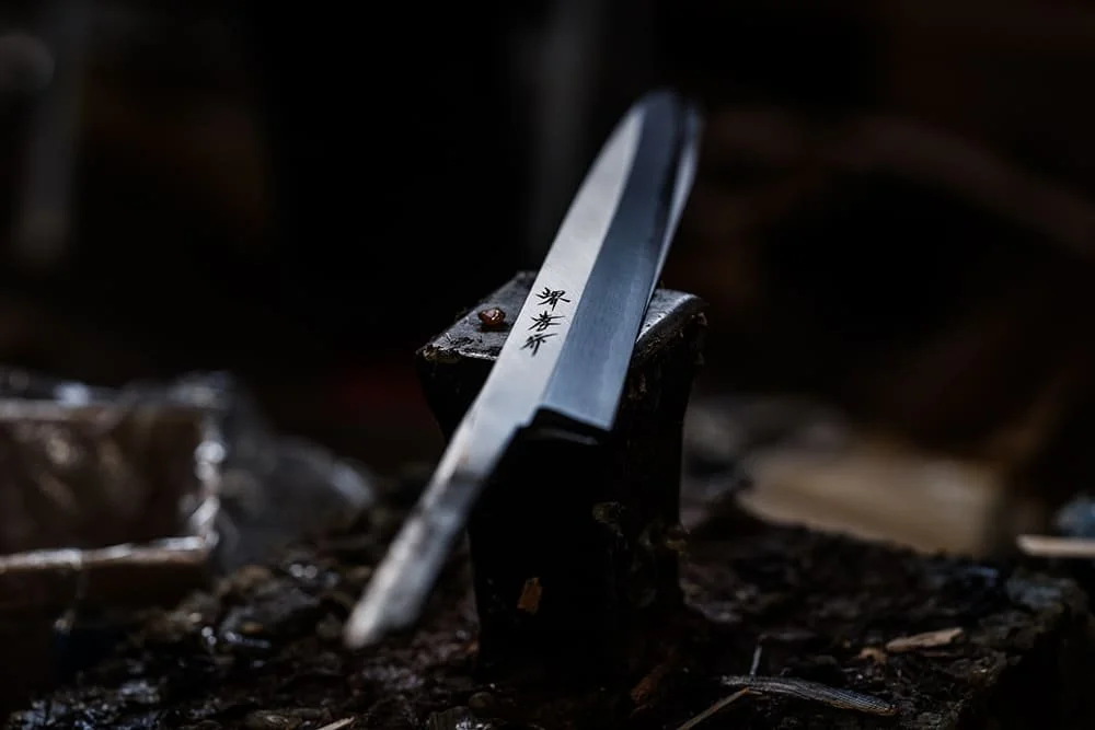 A Japanese kitchen knife resting on a small wooden block, with Japanese characters engraved on the blade, placed on a dark, textured surface.