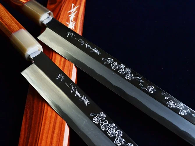 Two Japanese sushi knives with floral engravings on black blades, resting on a wooden and black surface.