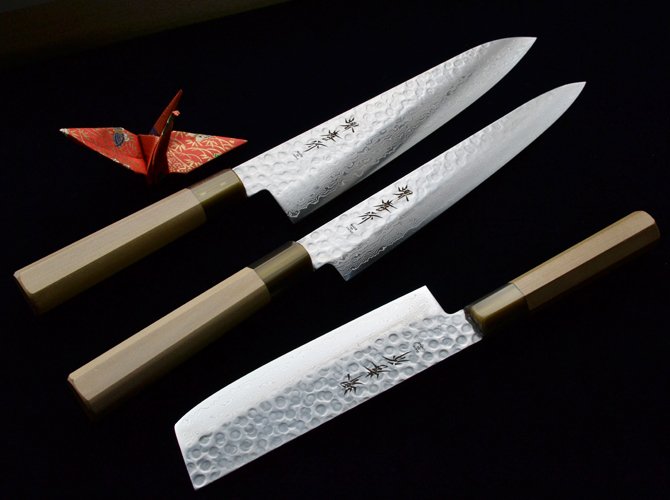 Three Japanese chef's knives with wooden handles and hammered pattern blades, accompanied by a paper origami crane on a black surface.