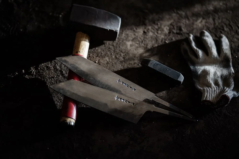 A hammer, two Japanese-style kitchen knives, a sharpening stone, and a work glove on a dark work surface.