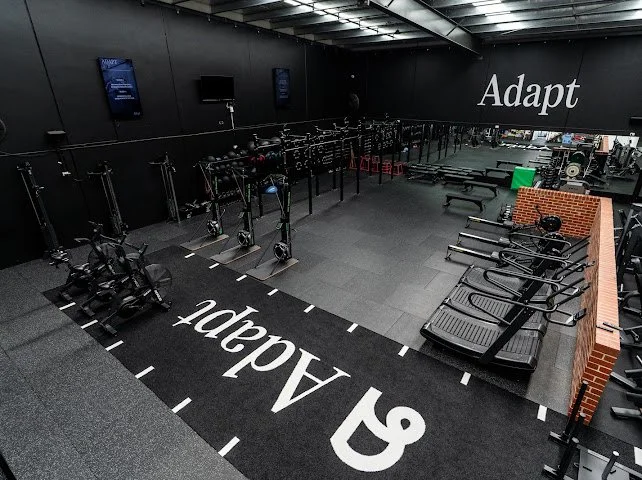 Empty gym with workout equipment, treadmills, rowing machines, and a brick wall. Black walls with the word 'Adapt' displayed prominently.