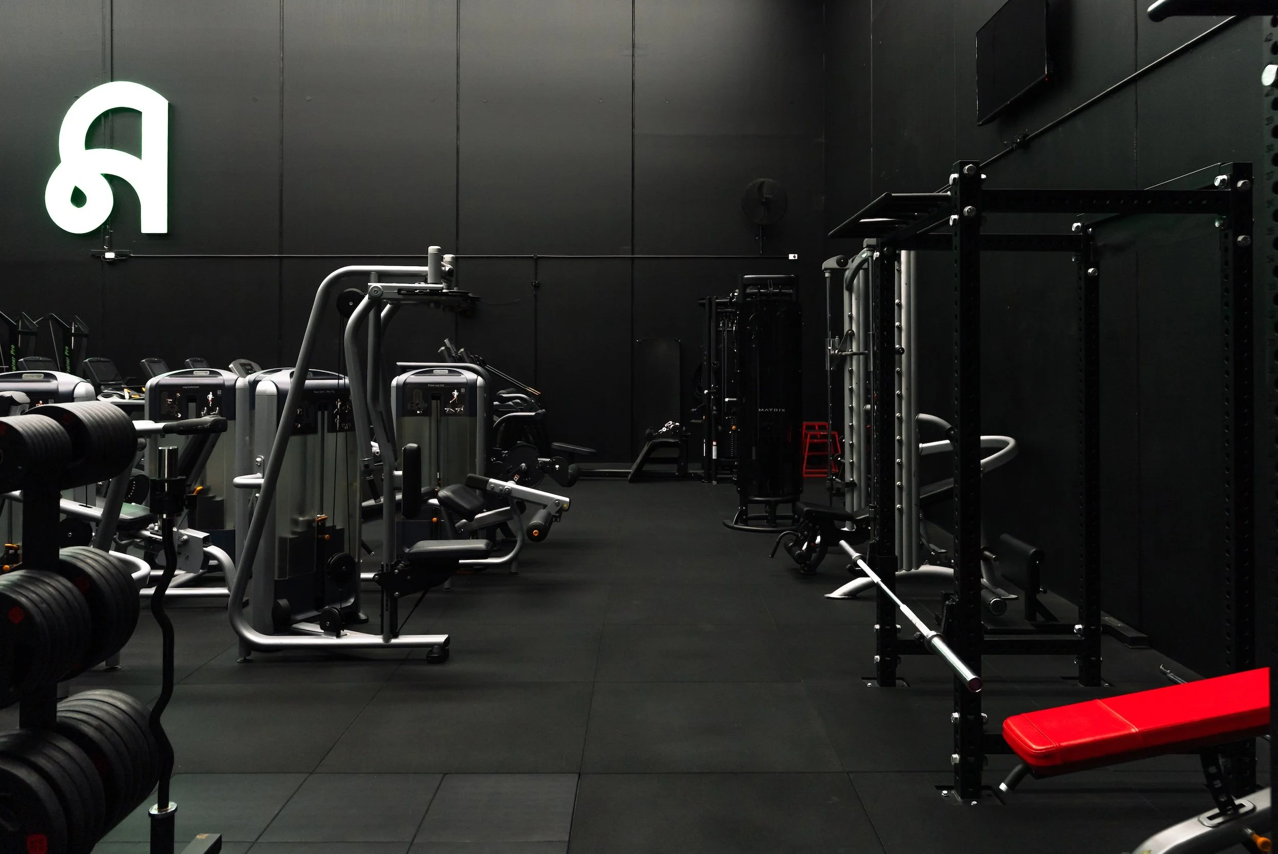 Inside a black gym with various workout equipment including weight machines, free weights, and a red workout bench.