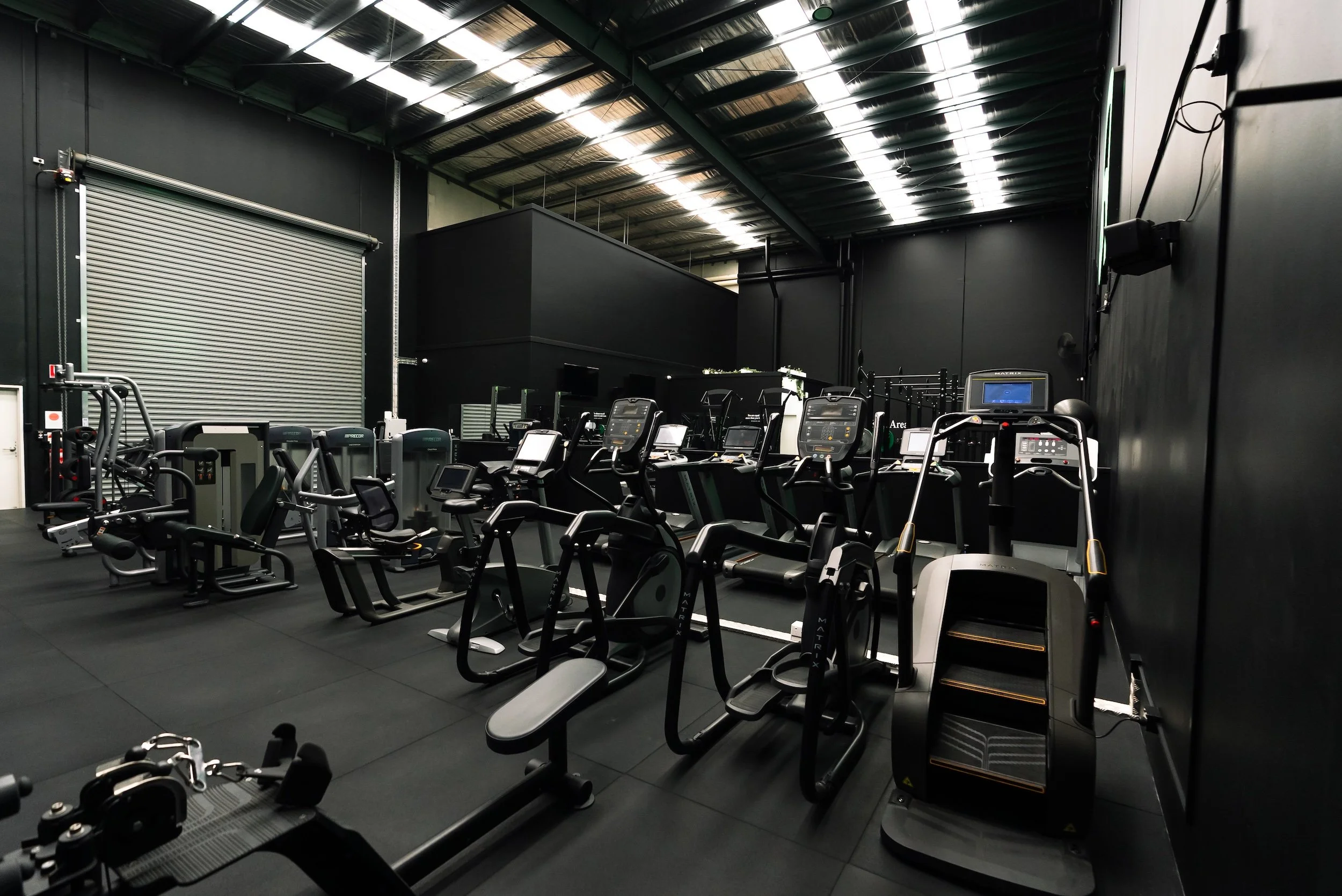Empty gym with black walls, ceiling, and flooring, featuring various exercise machines including treadmills and ellipticals, with a large metal rolling door on the left and industrial lighting.