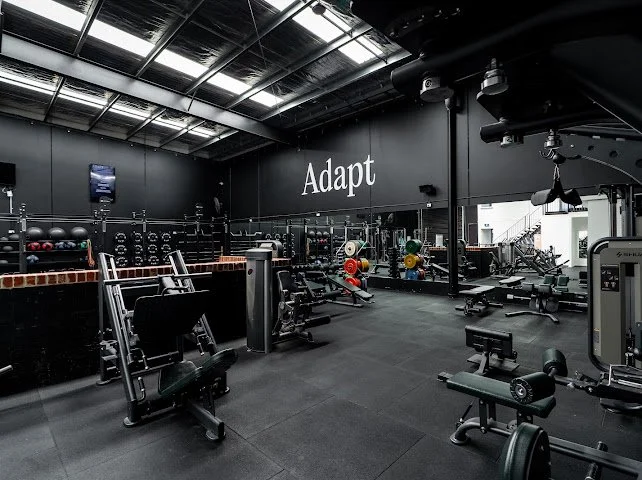 Empty gym with various workout equipment and motivational word 'Adapt' on the wall.