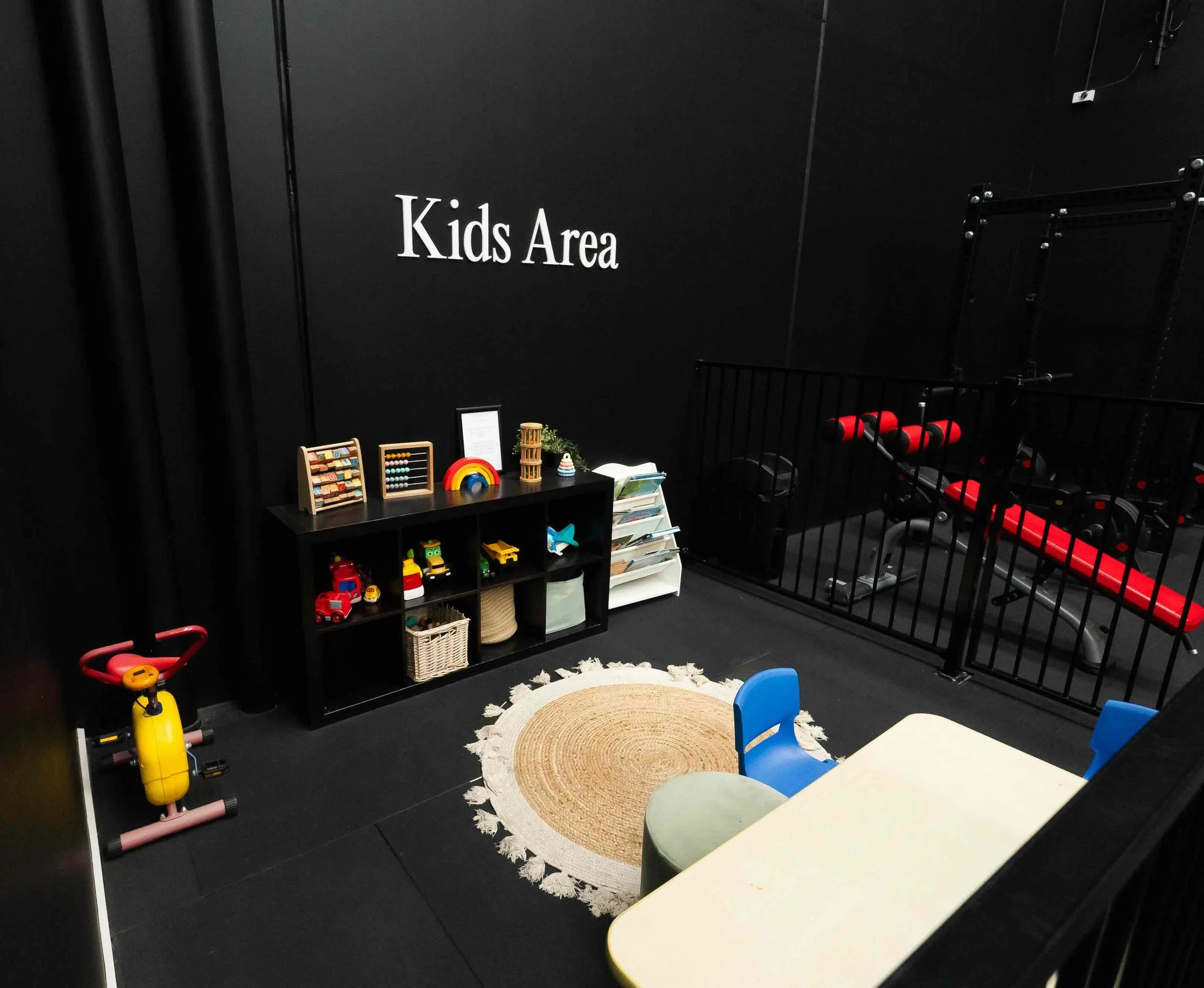 A children's play area with a black wall labeled 'Kids Area', featuring small tables and chairs, a black shelf with toys, colorful decorations, and black fencing.