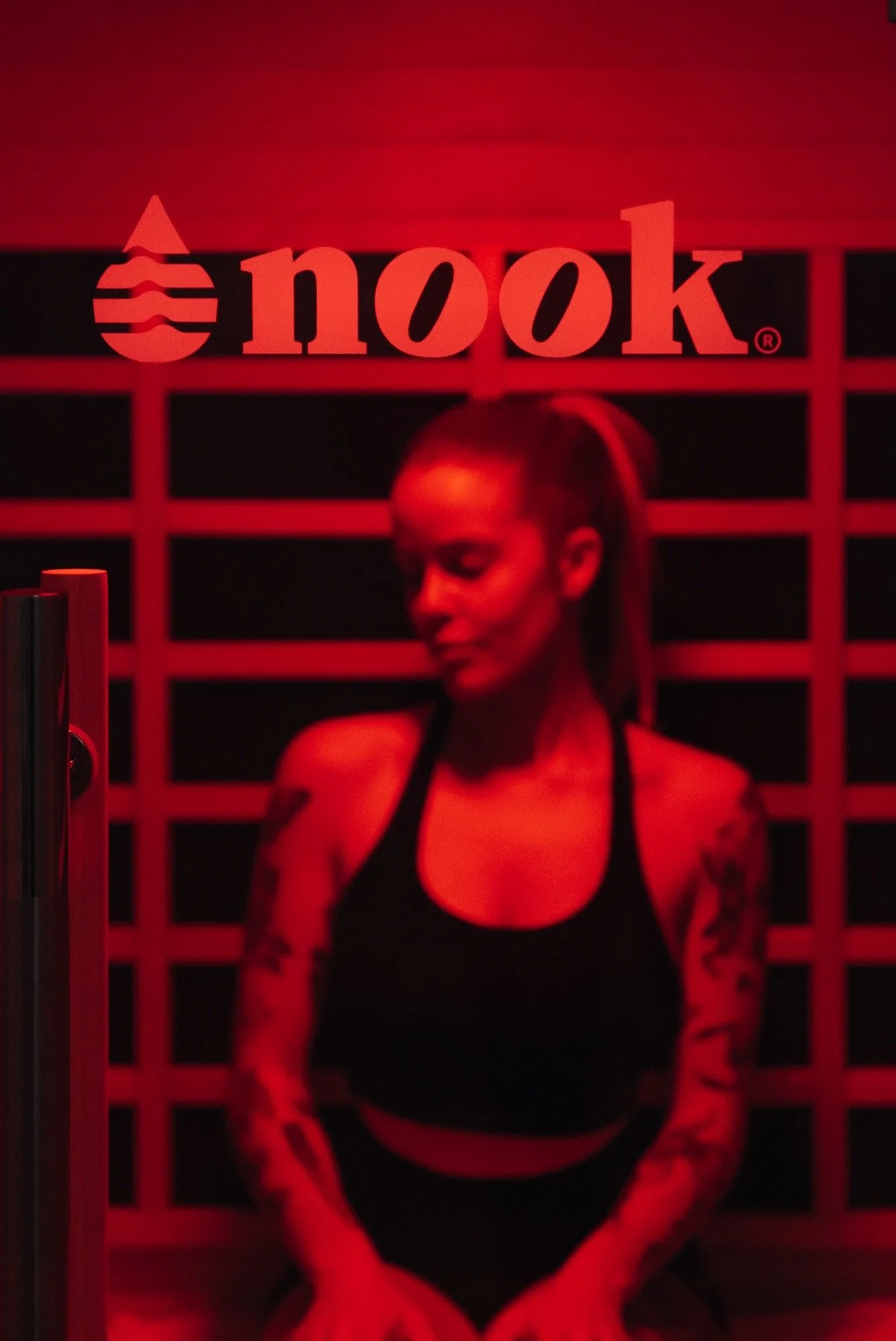 A woman with tattoos on her arms and dark hair tied back, hunching slightly with her eyes closed, in a dimly lit room with red lighting, a grid pattern on the wall, and a sign that reads 'nook' with a logo.