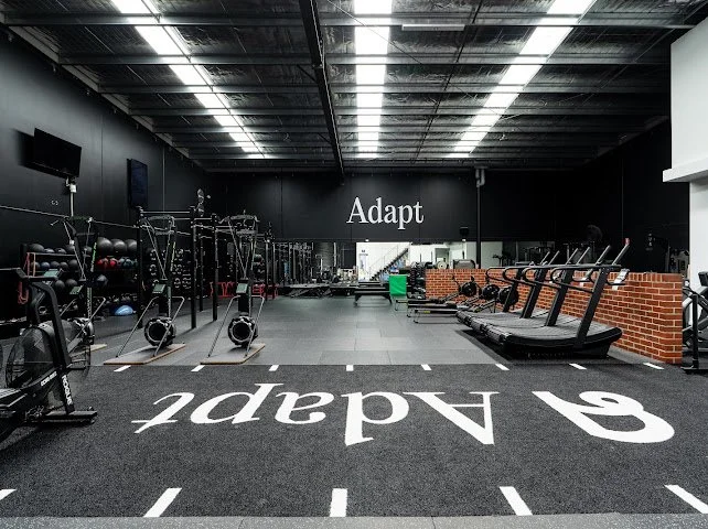 Empty gym with workout equipment, treadmills, weights, and a large sign that says 'Adapt' on the back wall.