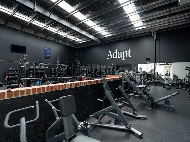 Empty gym with workout machines, weights, and a black wall with the word 'Adapt' printed on it.