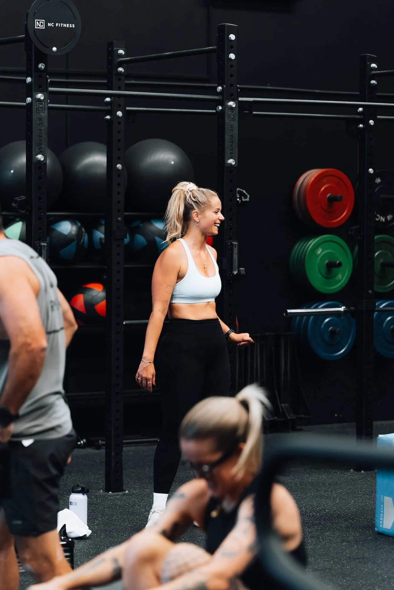 Woman in a white sports bra and black leggings smiling during a workout session at a gym, with various exercise equipment and other people visible around her.