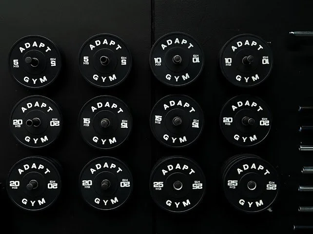 Set of black ADAPT GYM weight plates with white markings in various weights, arranged on a black rack.