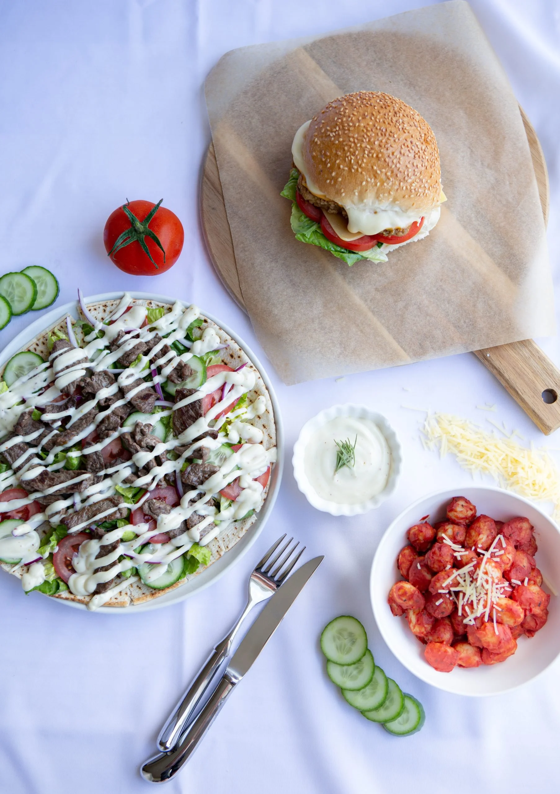 A table set with various dishes including a baked pizza topped with vegetables and meat, a bowl of spaghetti with tomato sauce and cheese, a bowl of meatballs, a burger with lettuce, tomato, cheese, and a sesame seed bun, a tomato, cucumber slices, and small bowls of cream and shredded cheese.