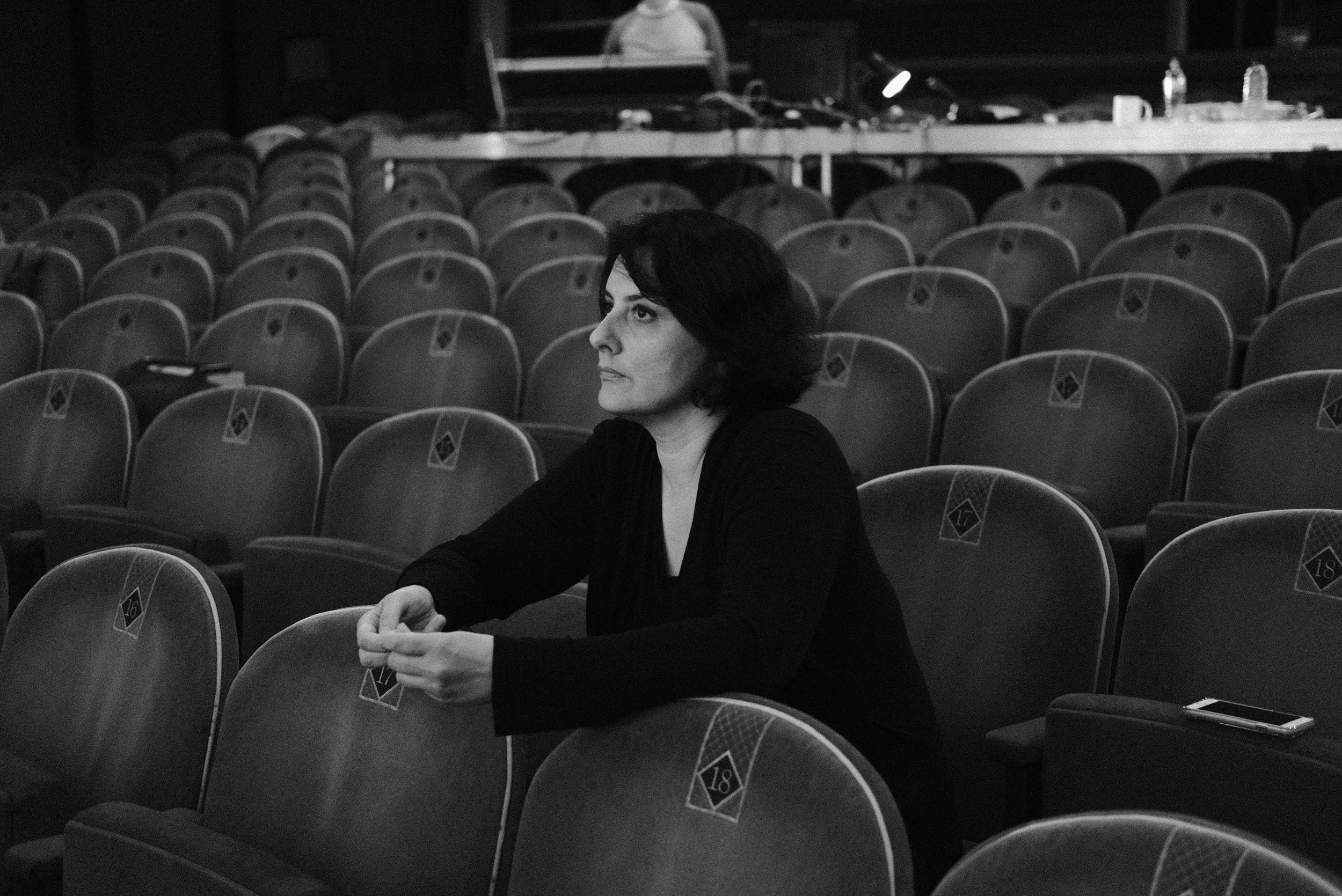 A woman with dark hair sitting alone in an empty theater or auditorium. She is looking towards the stage or screen, with her hands clasped together. The seats around her are unoccupied, and the theater appears dimly lit.