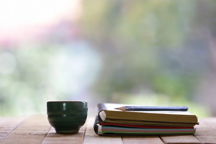 A dark green ceramic cup, a closed black pen, and a stack of books and folders are on a wooden table near a window with blurred outdoor scenery.
