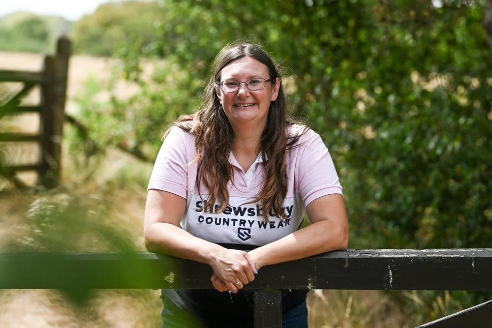 A woman with long brown hair and glasses leaning on a black wooden fence outdoors, smiling, with greenery and a rural landscape in the background.