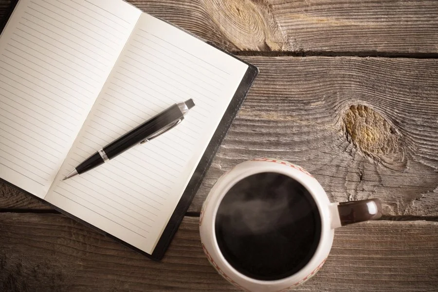 Open notebook with lined pages and a black pen on a wooden table next to a steaming cup of coffee.