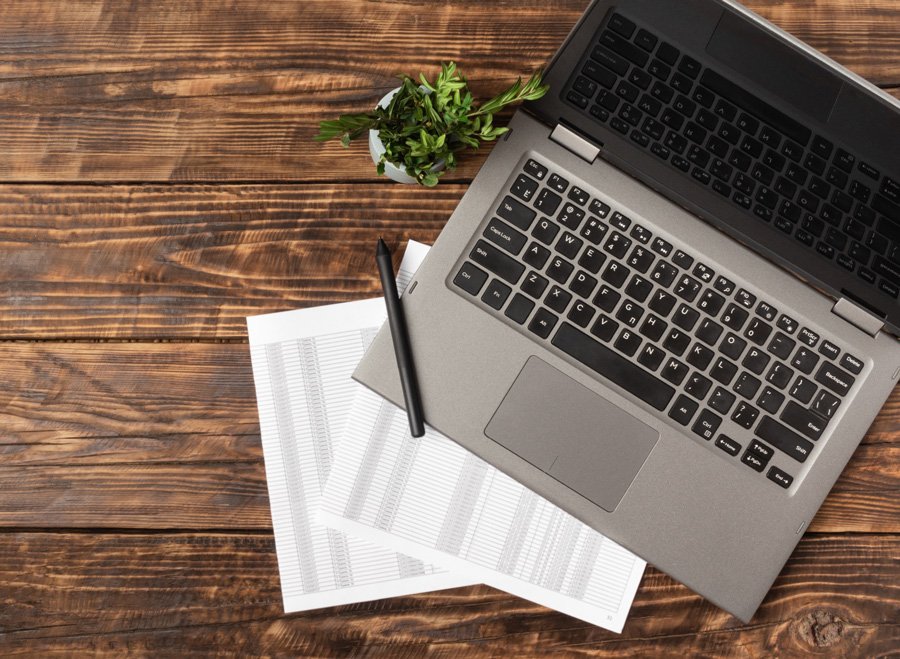 A laptop, a black pen, printed spreadsheets, and a small potted plant on a wooden desk.