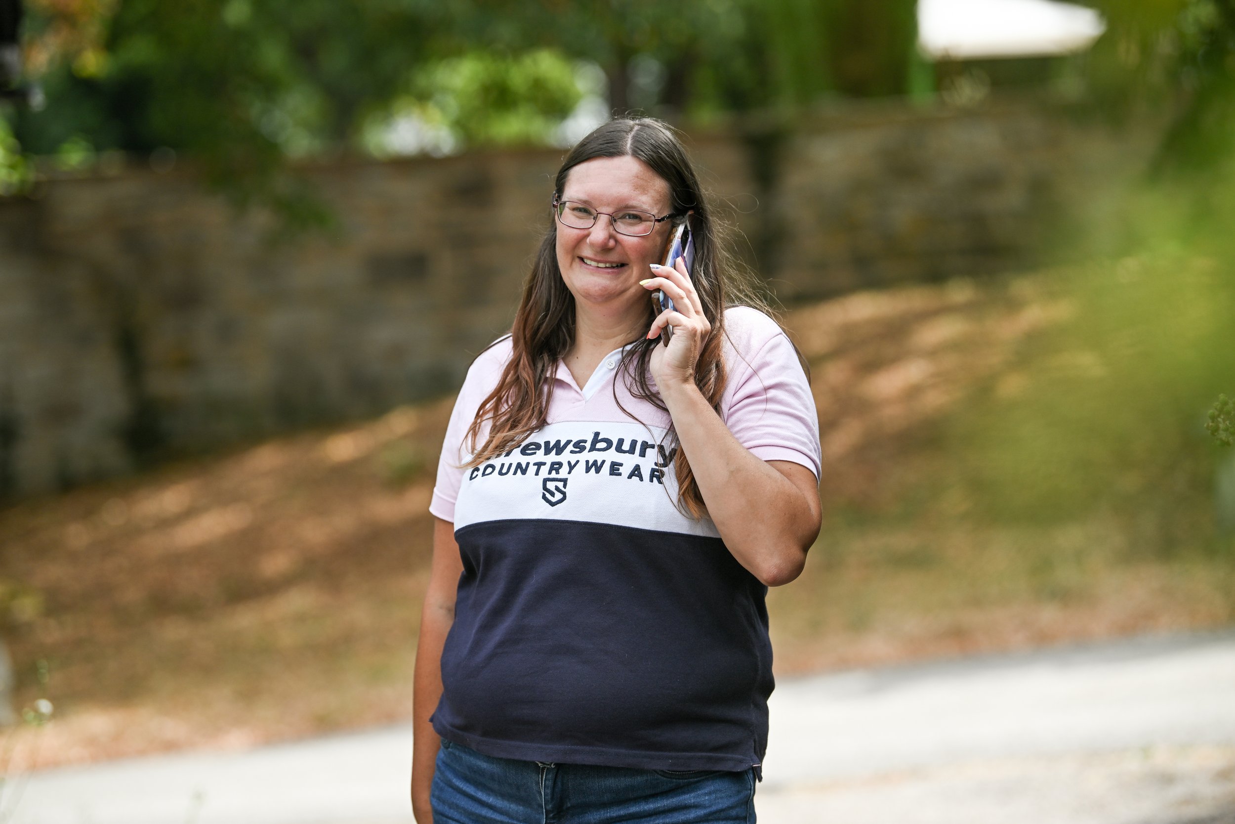 Woman with glasses and long hair smiling and talking on her phone outdoors.